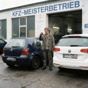 Ein frustrierter Mann steht mit den Schlüsseln eines ungenutzten Mietwagens neben seinem reparaturbedürftigen Wagen vor einer Werkstatt.