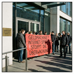 Ein großes, rot bemaltes Protestbanner blockiert den gläsernen Eingang einer modernen Geschäftsstelle; ein Demonstrant nutzt ein Megafon.